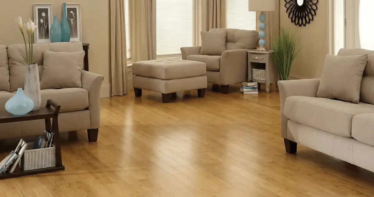 A living room with professionally installed bamboo flooring, beige sofas, a matching ottoman, a side table with a blue lamp, vases, framed pictures, and large windows with blinds and curtains in Tucson, AZ